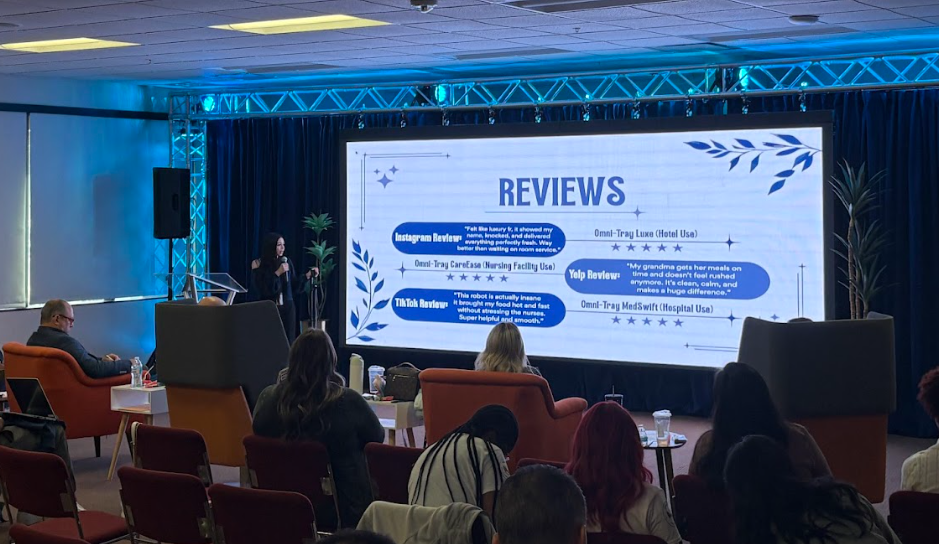 A high school student stands on a stage presenting an autonomous food delivery robot concept to a panel of judges during a business pitch competition.