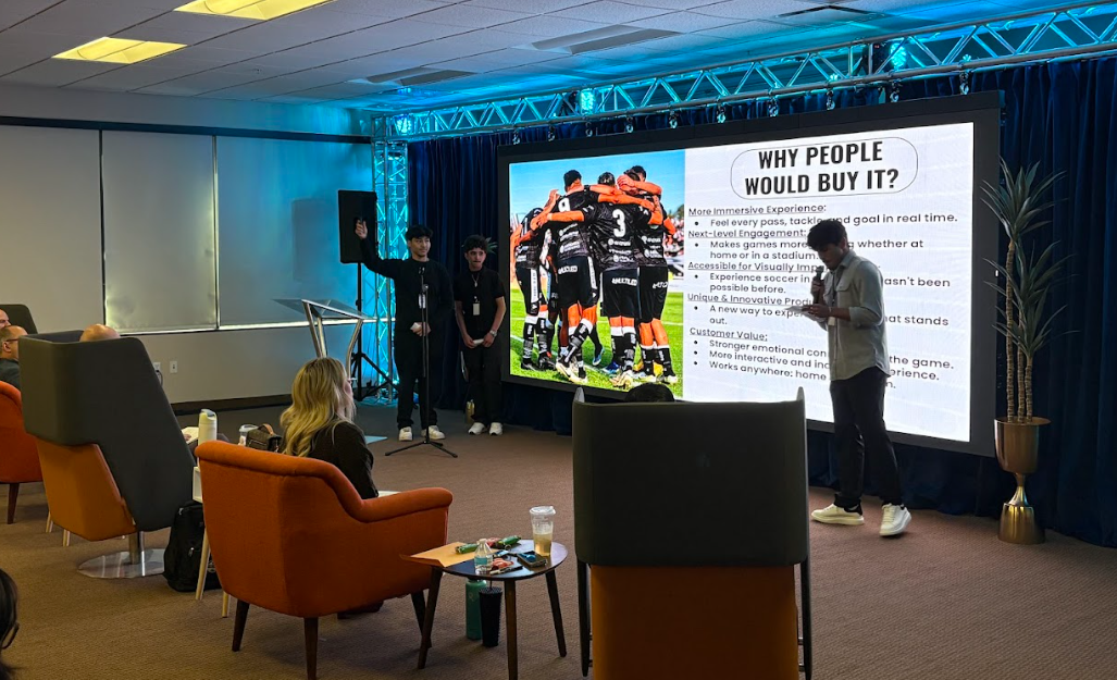 Three high school students present their invention, a device for visually impaired sports fans, to a panel of seated professional judges during a business pitch competition.